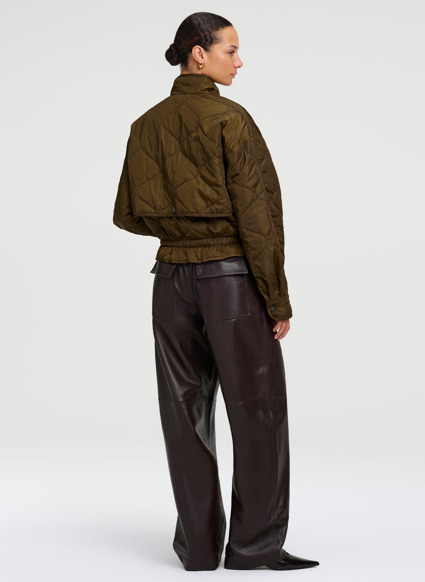 A model shown from the back wearing an olive green quilted cropped jacket and dark brown wide-leg leather trousers, photographed against a seamless light gray studio background.