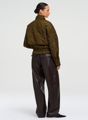 A model shown from the back wearing an olive green quilted cropped jacket and dark brown wide-leg leather trousers, photographed against a seamless light gray studio background.