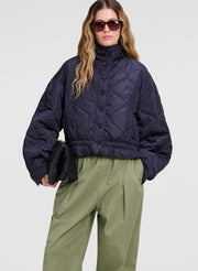 A model wearing a cropped, quilted navy blue jacket with a high collar and button front, paired with olive green trousers and dark sunglasses, against a light gray studio background.