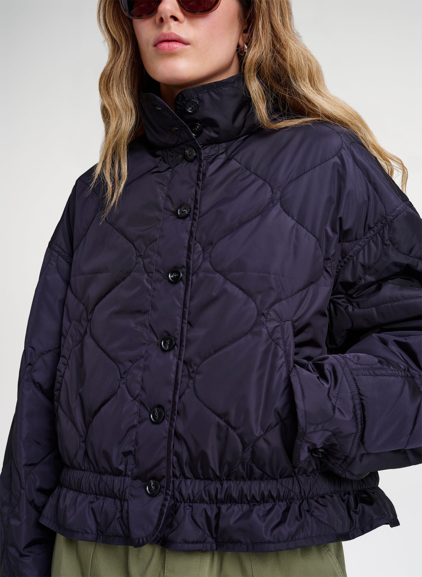 A close-up of a model wearing a dark navy blue quilted puffer jacket with a high stand-up collar and front button closure, paired with olive green bottoms against a light gray studio background.