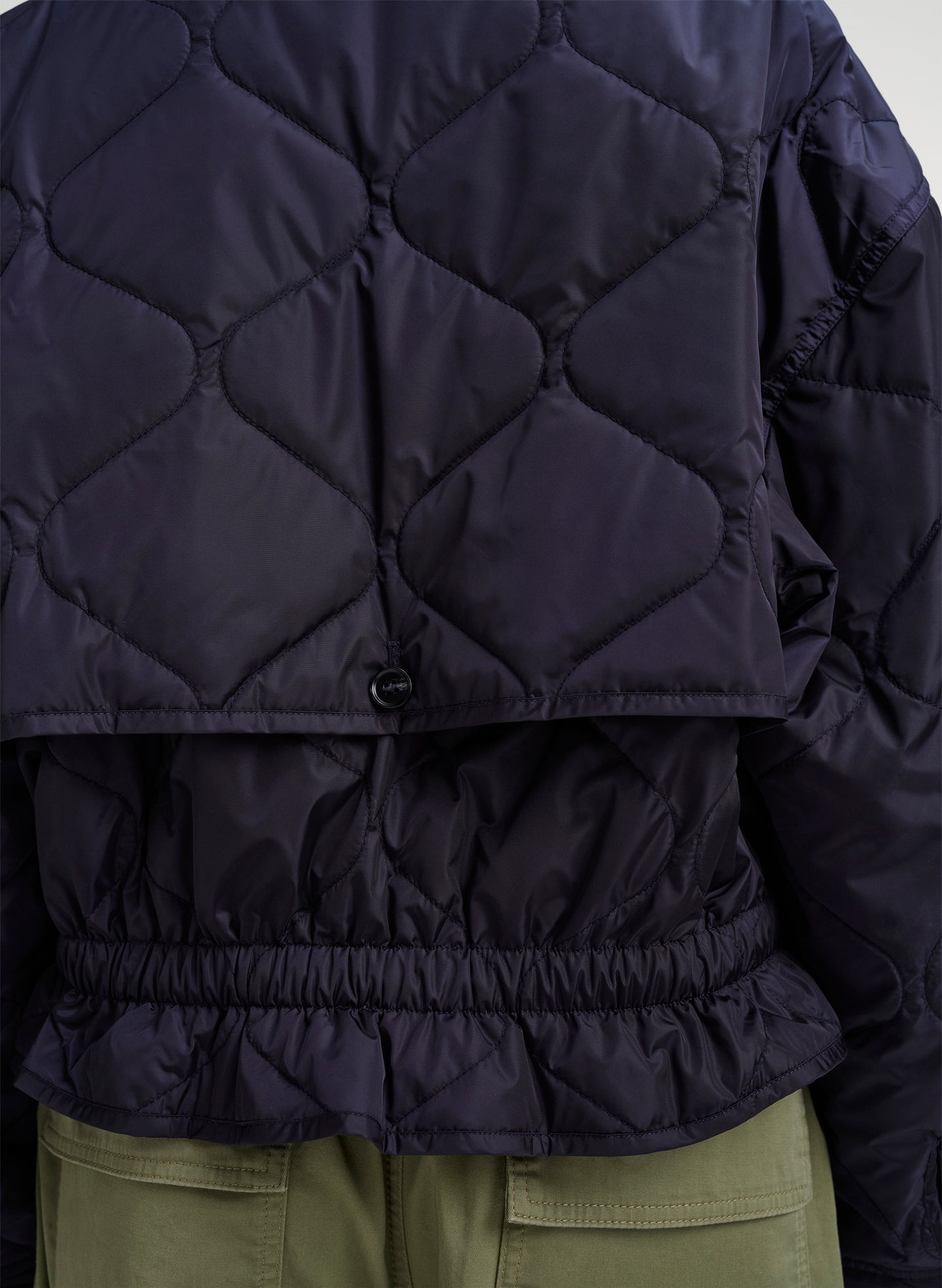 Close-up of the back of a dark navy quilted jacket with a diamond pattern, elasticated hem, and a small button closure, worn over olive green pants.