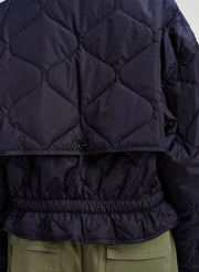 Close-up of the back of a dark navy quilted jacket with a diamond pattern, elasticated hem, and a small button closure, worn over olive green pants.