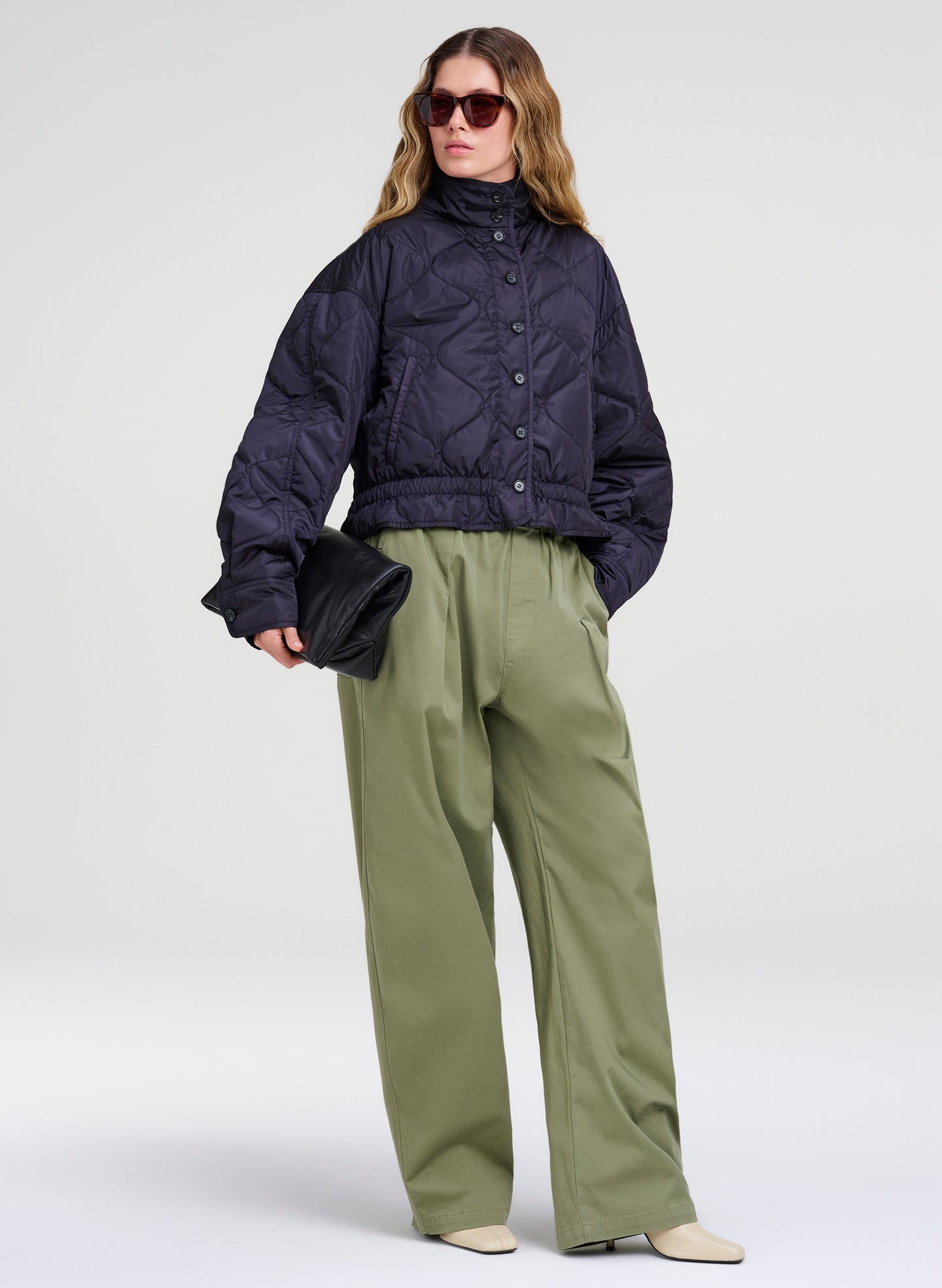 A model wearing a cropped, dark navy blue quilted puffer jacket, olive green wide-leg trousers, and beige pointed-toe flats, holding a black clutch against a light gray studio background.