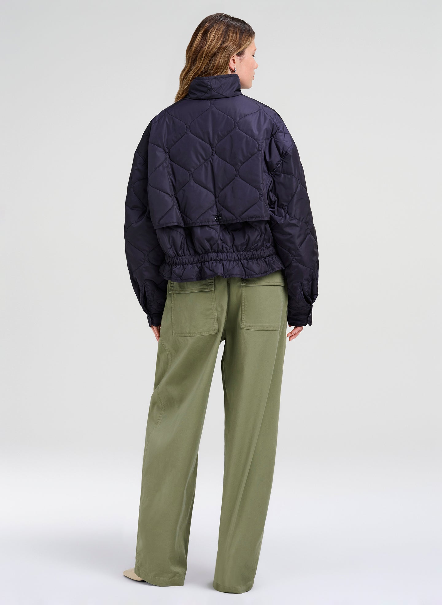 A model shown from the back wearing a dark navy quilted puffer jacket and olive green trousers, photographed against a neutral light gray studio background.