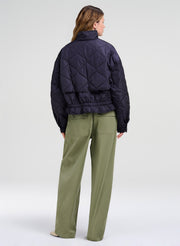 A model shown from the back wearing a dark navy quilted puffer jacket and olive green trousers, photographed against a neutral light gray studio background.