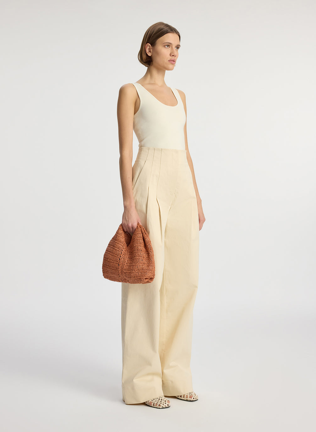 front view of woman wearing beige tank top and beige wide leg pants carrying small raffia woven handbag in brown