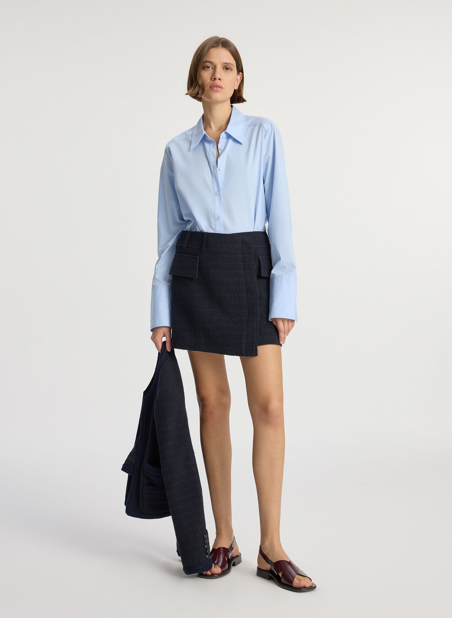 front view of woman wearing blue button down collared shirt and navy tweed mini wrap skirt while holding jacket