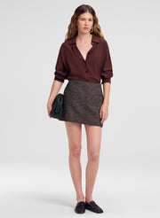 A model stands against a light gray studio background wearing a dark brown button-up shirt, a textured gray mini skirt, black loafers, and holding a dark green textured clutch.
