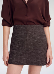 A model wearing a dark brown textured tweed mini skirt paired with a dark brown silk blouse, shown from the chest down against a light gray studio background.