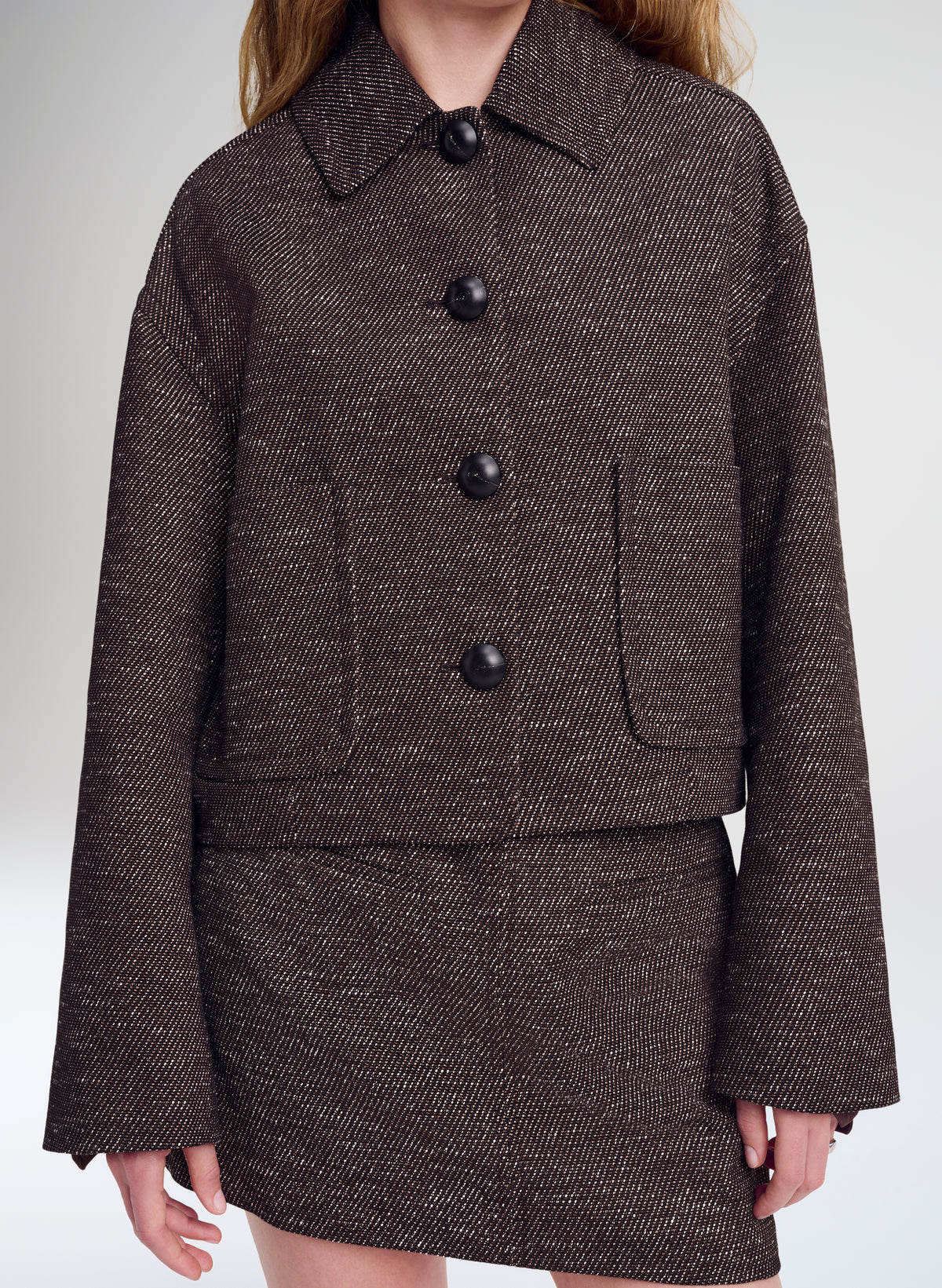 A cropped, long-sleeve tweed jacket in a dark brown and black herringbone pattern, featuring a pointed collar, four large black buttons, and two front patch pockets, shown on a model against a light gray studio background.