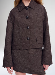 A cropped, long-sleeve tweed jacket in a dark brown and black herringbone pattern, featuring a pointed collar, four large black buttons, and two front patch pockets, shown on a model against a light gray studio background.