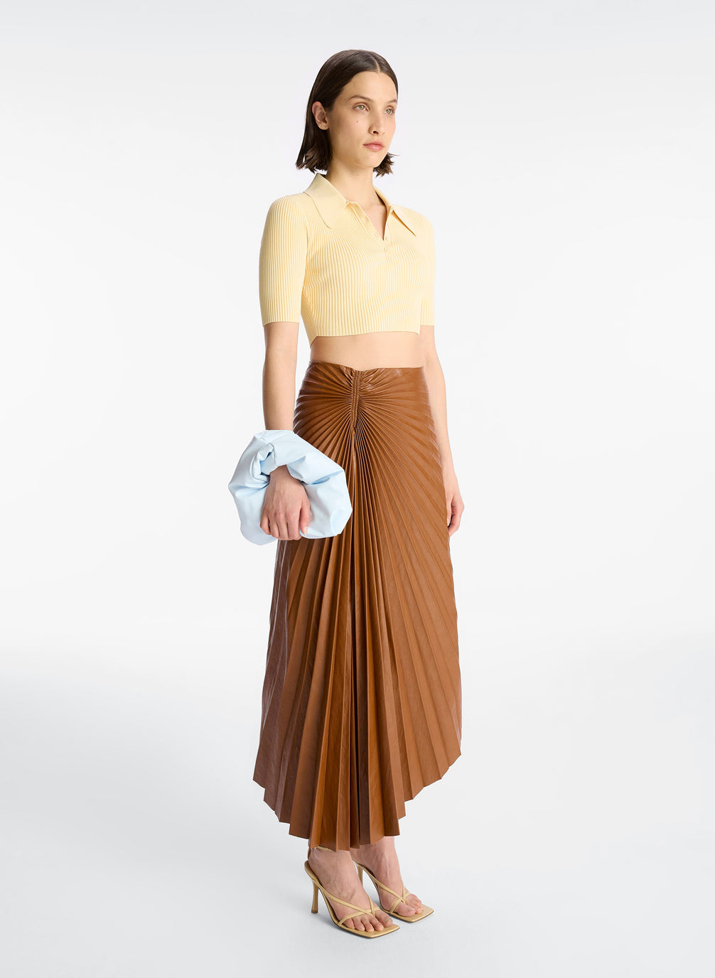 side view of woman wearing cream cropped collared knit shirt and brown pleated vegan leather skirt