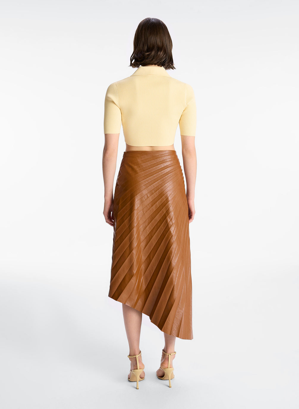 back view of woman wearing cream cropped collared knit shirt and brown pleated vegan leather skirt
