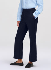 A model wearing high-waisted, dark navy blue cropped trousers, styled with a light blue button-down shirt and black loafers, against a plain light gray studio background.