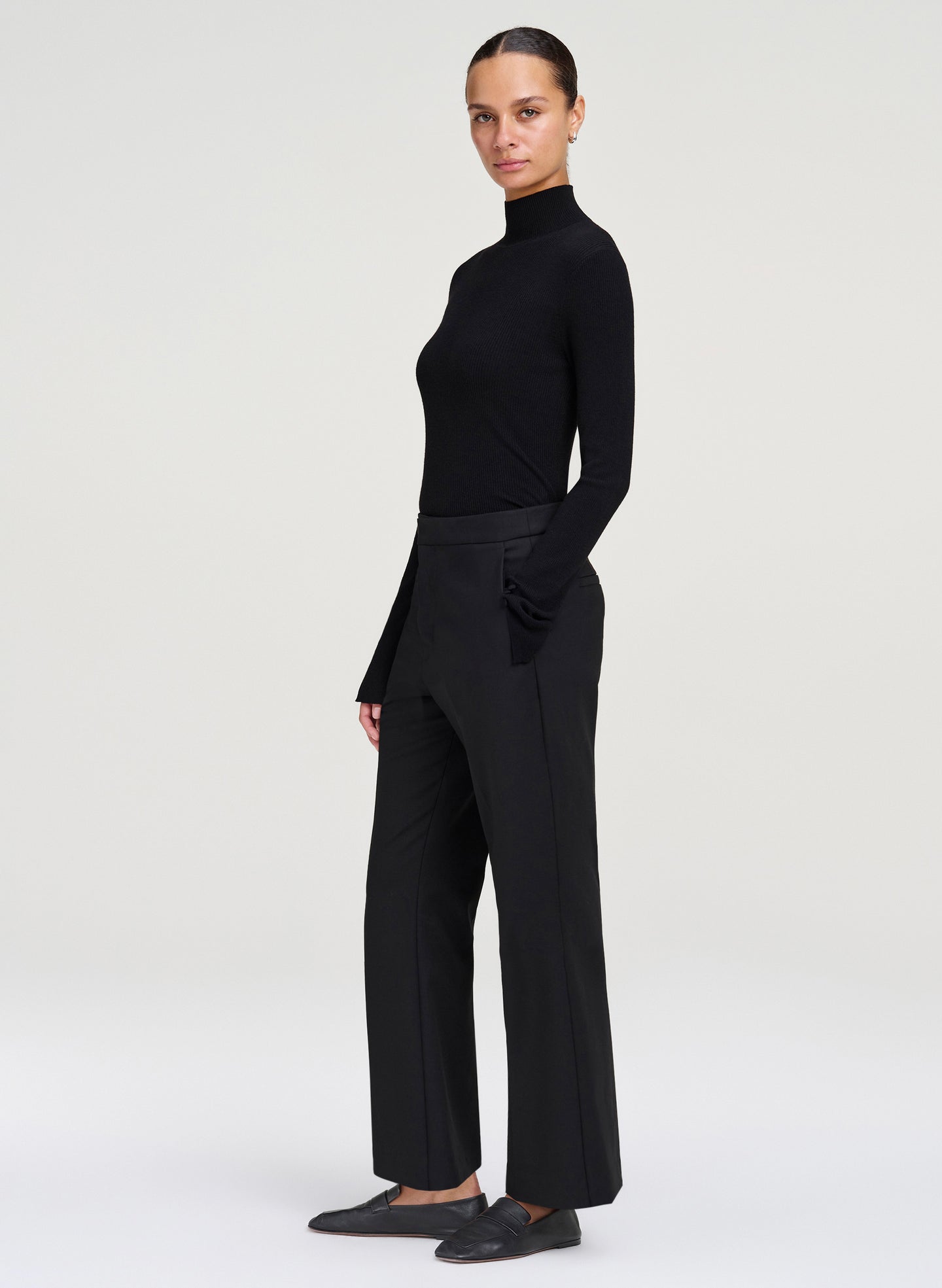 A model wearing a black long-sleeve mock neck top and matching black wide-leg trousers, paired with black slip-on shoes, standing against a light gray studio background.