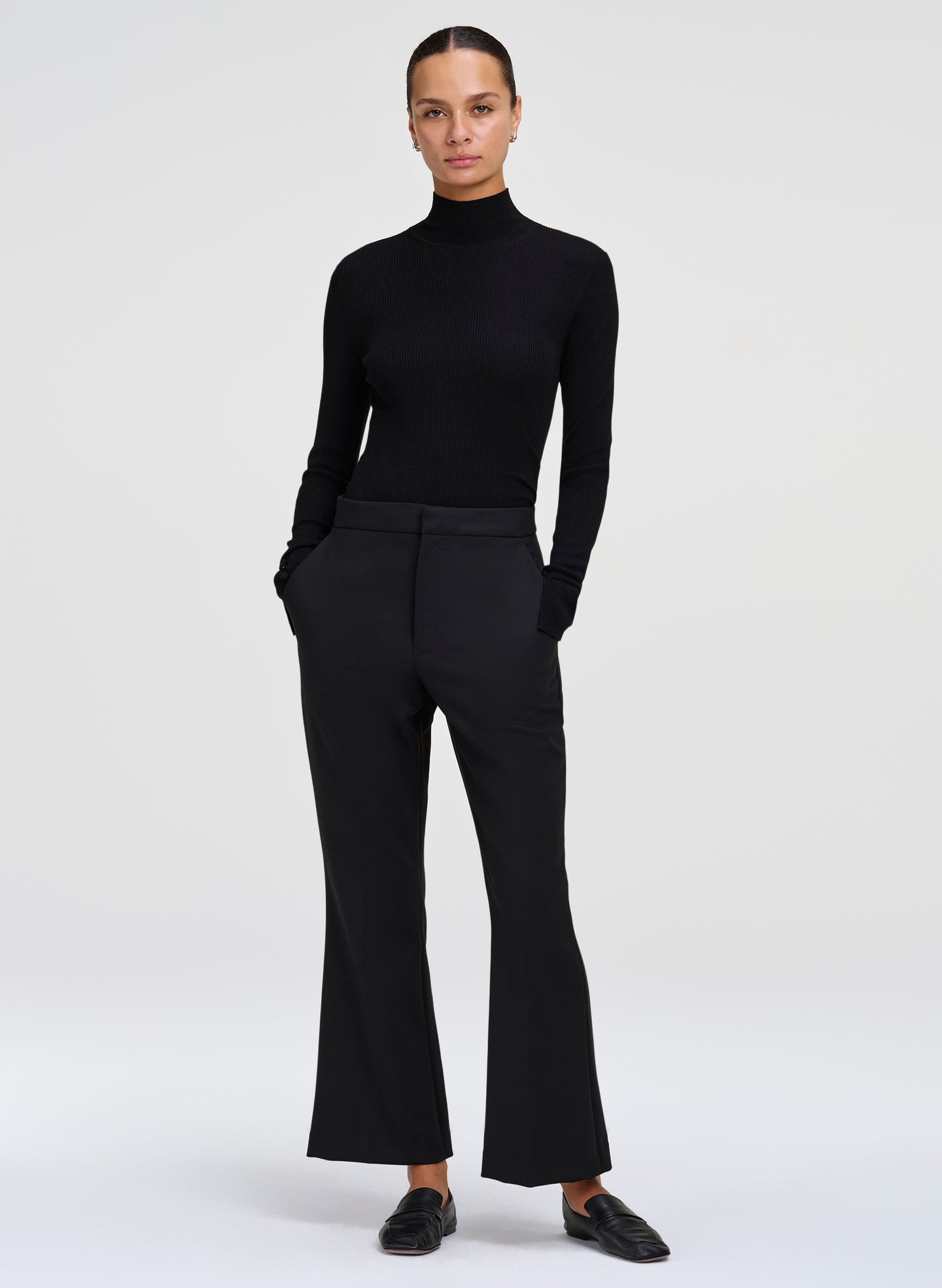 A model wearing a black long-sleeve mock neck top and matching black flared trousers, paired with black loafers, against a light gray studio background.