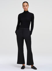A model wearing a black long-sleeve mock neck top and matching black flared trousers, paired with black loafers, against a light gray studio background.
