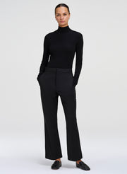 A model wearing a black long-sleeve mock neck top and matching black wide-leg trousers, paired with black loafers, against a light gray studio background.