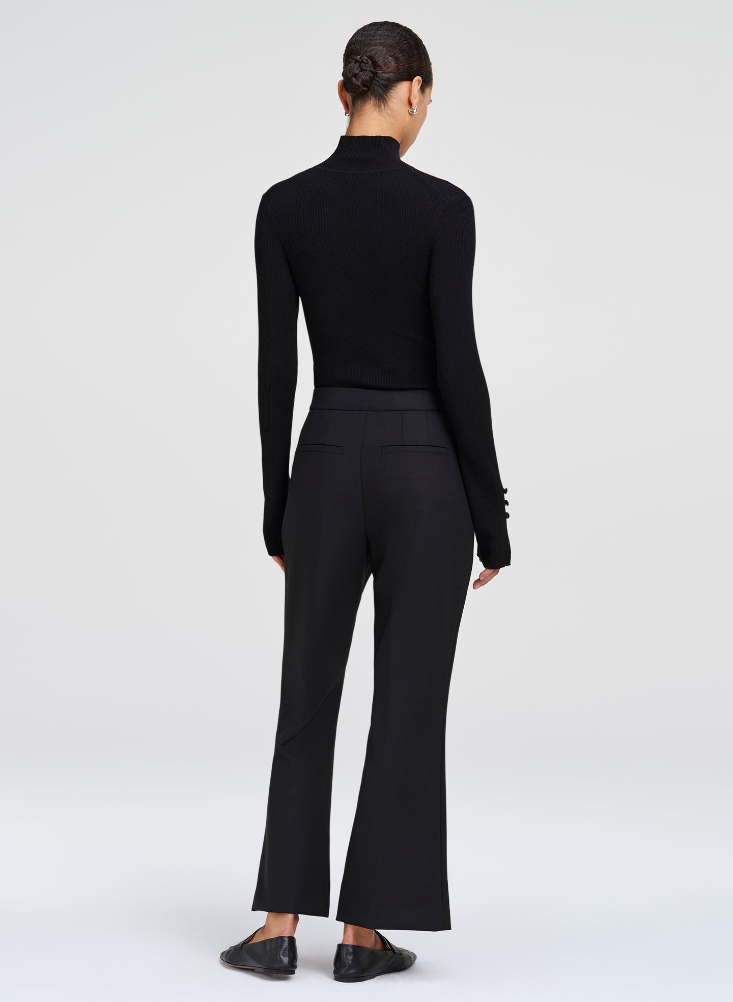 A model shown from the back wearing a black long-sleeve mock neck top and matching black flared trousers, paired with black flats, against a light gray studio background.