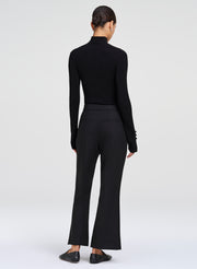 A model shown from the back wearing a black long-sleeve mock neck top and matching black flared trousers, paired with black flats, against a light gray studio background.