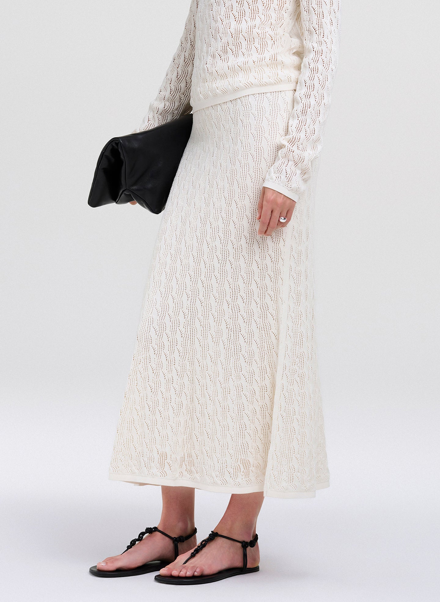 A model wearing a long-sleeve cream lace midi skirt with a matching cream lace top, holding a black clutch, and wearing black strappy flat sandals against a light gray studio background.