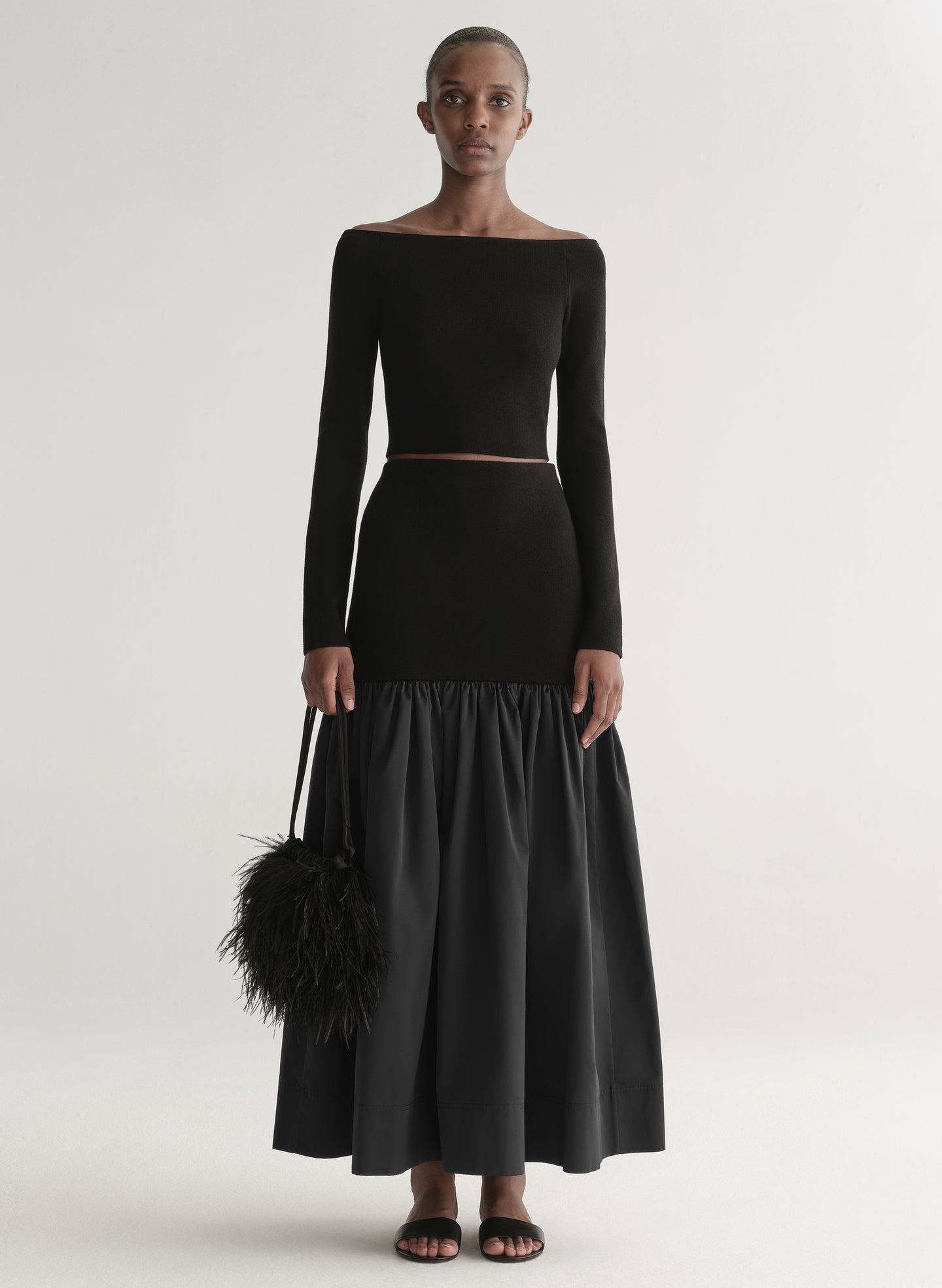 Woman wearing a black off-shoulder top and long skirt with a black feathered bag on a white background