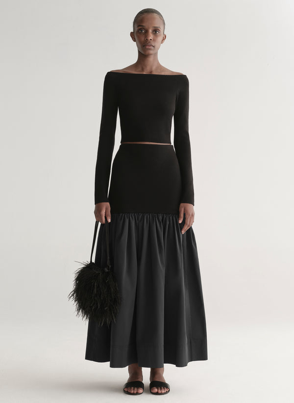 Woman wearing a black off-shoulder top and long skirt with a black feathered bag on a white background