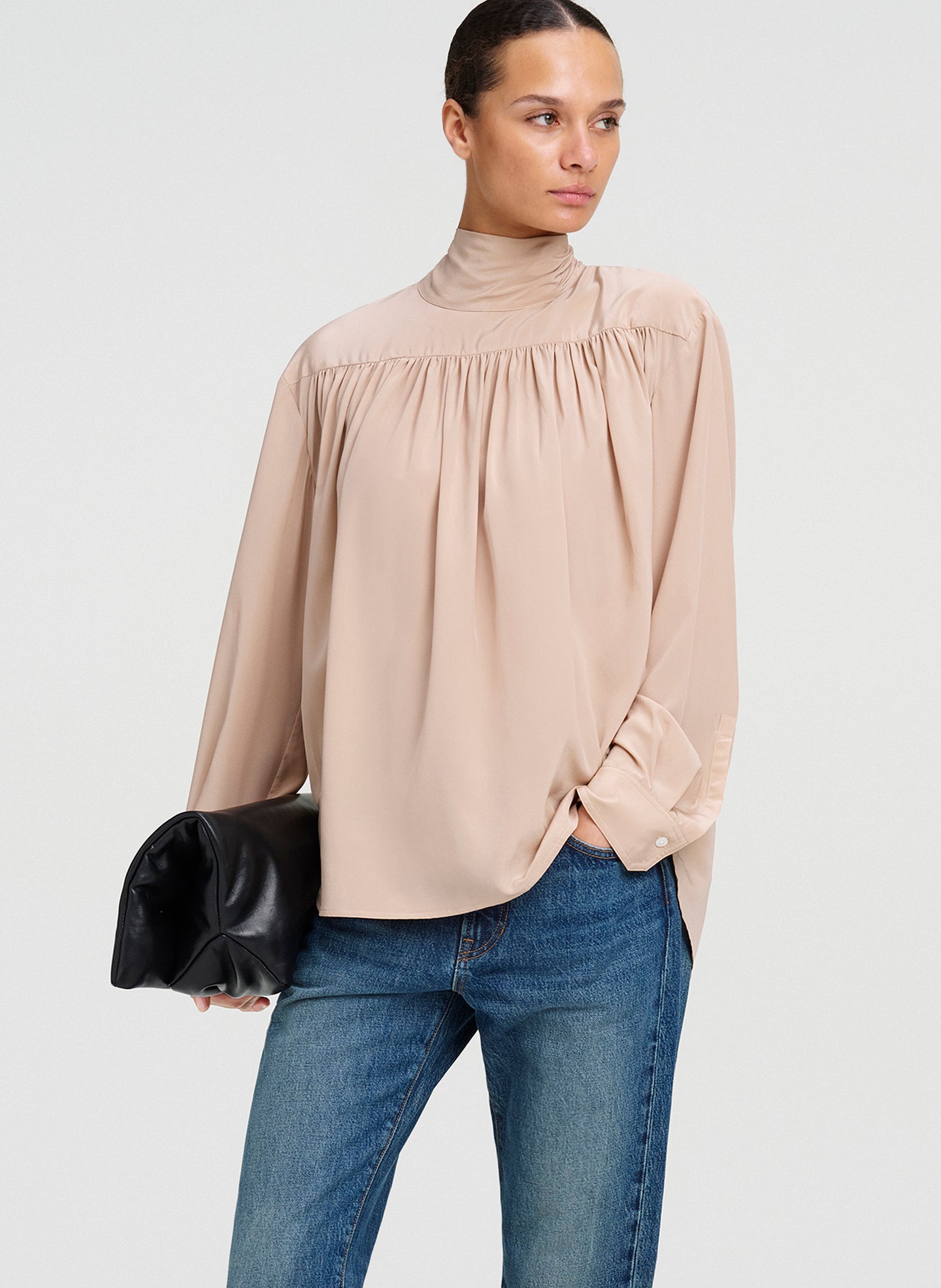 A woman modeling a beige high-neck long-sleeve blouse with gathered detailing at the chest, paired with blue jeans and holding a black clutch bag, against a light gray studio background.