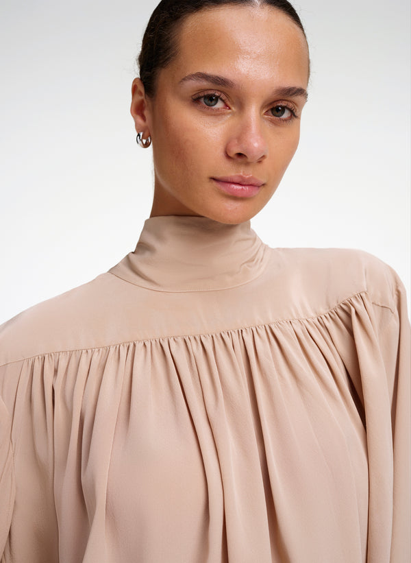 A close-up of a model wearing a beige high-neck blouse with gathered, pleated detailing across the chest and shoulders.