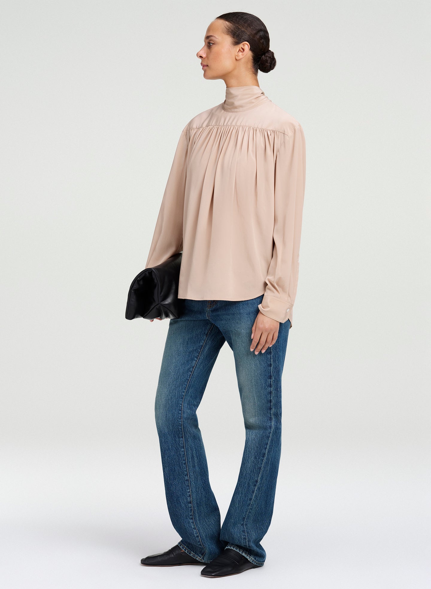 A woman modeling a light beige high-neck long-sleeve blouse with gathered detailing at the chest, paired with blue jeans and black shoes, holding a black clutch against a plain light gray studio background.