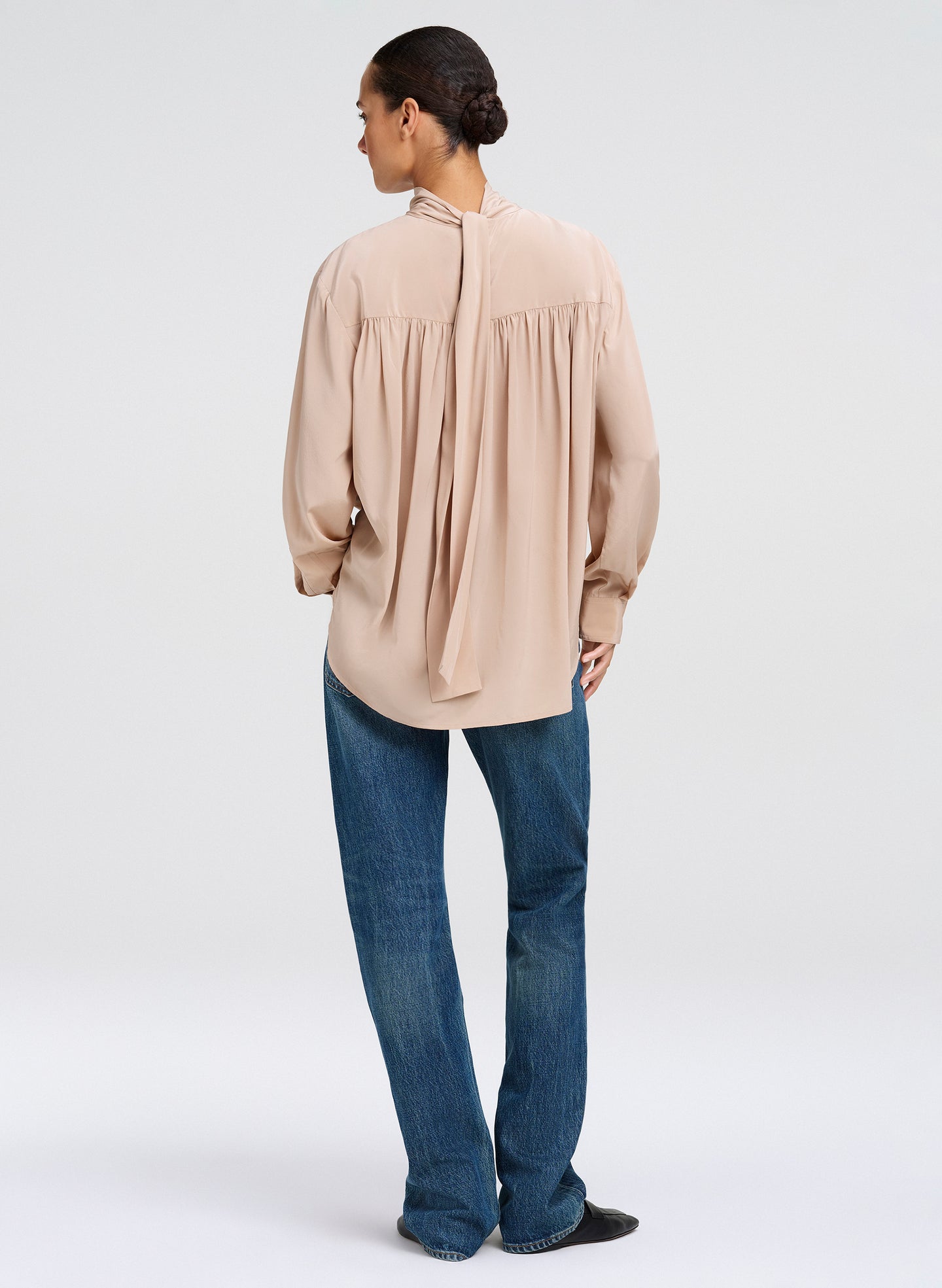 A woman shown from the back wearing a light beige long-sleeve blouse with a gathered back and tie detail, paired with blue jeans, against a plain light gray studio background.