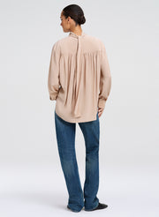 A woman shown from the back wearing a light beige long-sleeve blouse with a gathered back and tie detail, paired with blue jeans, against a plain light gray studio background.