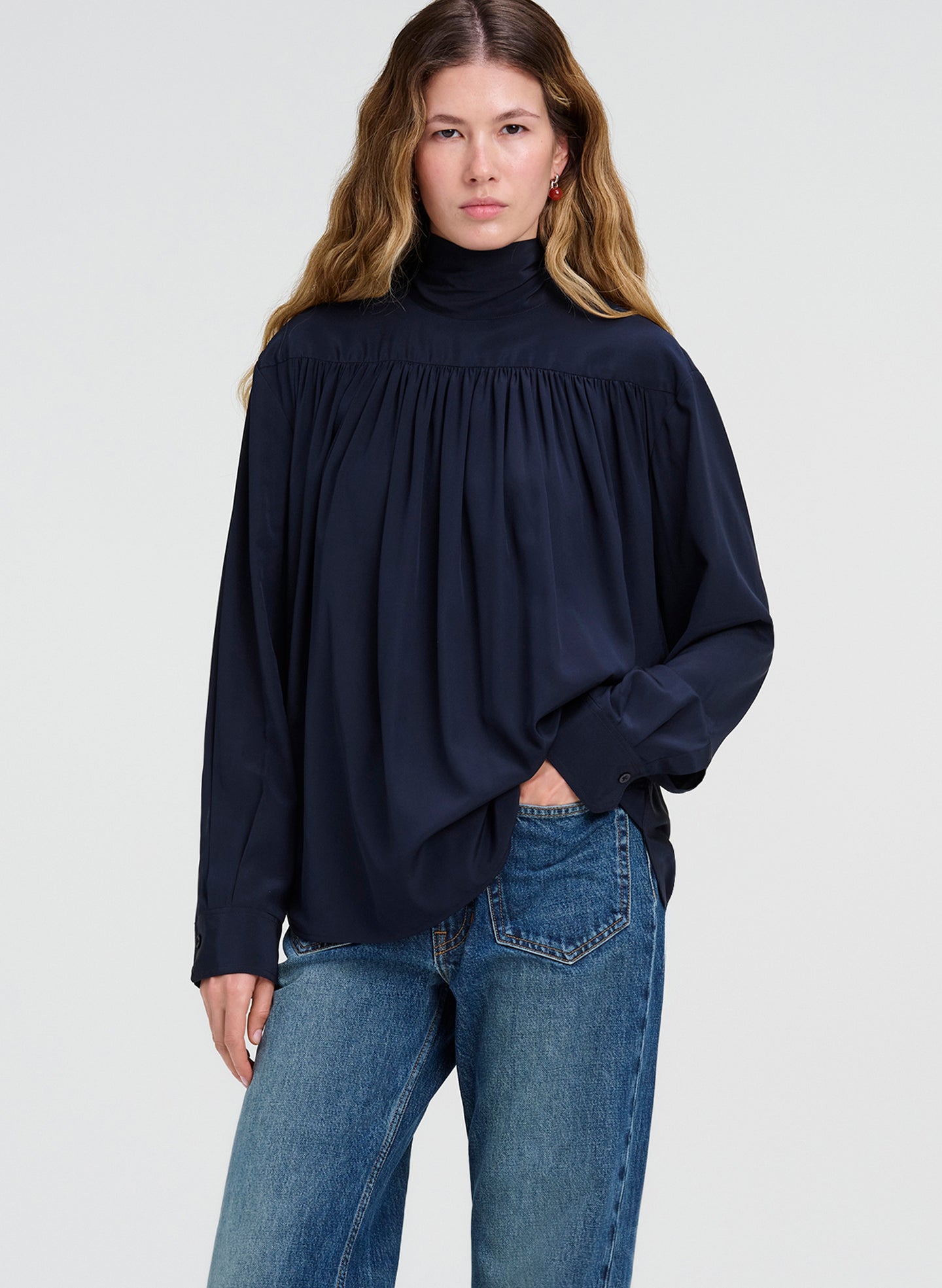 A woman modeling a dark navy blue long-sleeve blouse with a gathered smocked neckline, paired with blue jeans, against a light gray studio background.