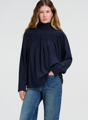 A woman modeling a dark navy blue long-sleeve blouse with a gathered smocked neckline, paired with blue jeans, against a light gray studio background.
