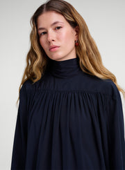 A woman with long light brown hair wearing a dark navy blue high-neck blouse with a gathered, pleated bodice and wide sleeves, set against a plain light gray studio background.