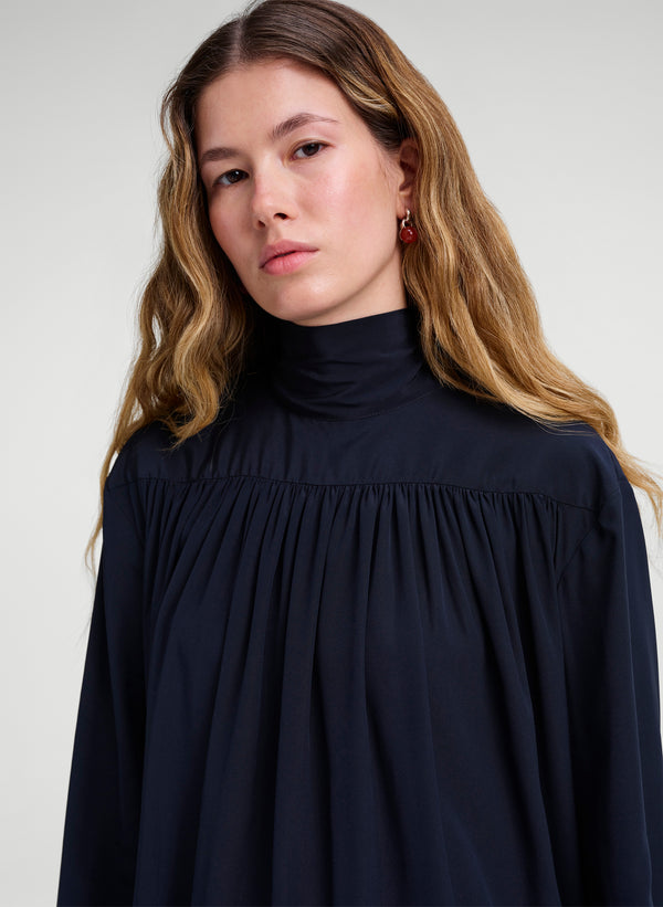 A woman with long light brown hair wearing a dark navy blue high-neck blouse with a gathered, pleated bodice and wide sleeves, set against a plain light gray studio background.