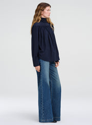 A woman modeling a dark navy blue long-sleeve blouse with a high gathered neckline and wide-leg blue jeans against a plain light gray studio background.