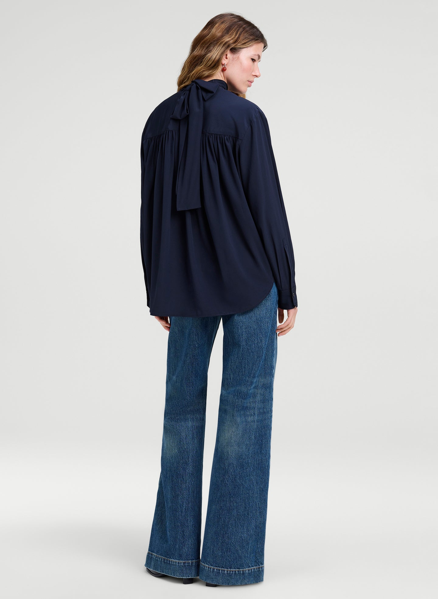 A woman shown from the back wearing a navy blue long-sleeve blouse with a large bow detail at the back neckline and blue flared jeans, photographed against a light gray studio background.
