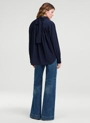 A woman shown from the back wearing a navy blue long-sleeve blouse with a large bow detail at the back neckline and blue flared jeans, photographed against a light gray studio background.