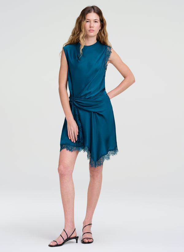 A woman modeling a sleeveless teal wrap-style dress with lace trim and black strappy high-heeled sandals against a light gray studio background.