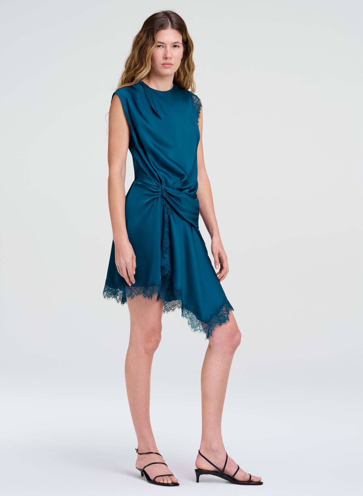 A woman modeling a sleeveless teal blue dress with a gathered twist detail at the waist and a lace hem, paired with black strappy heels, against a light gray studio background.