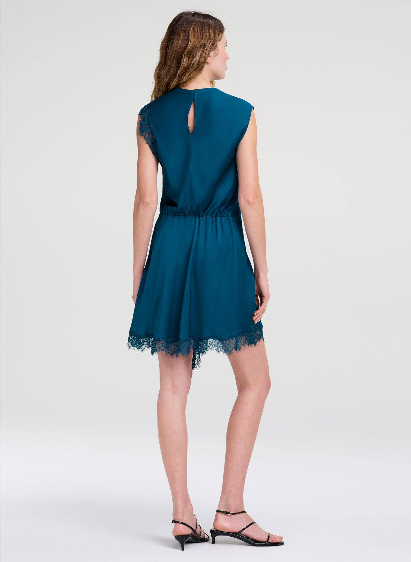 A woman shown from the back wearing a short teal blue dress with a lace hem and keyhole back detail, paired with black strappy heels, against a light gray studio background.