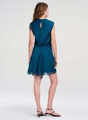A woman shown from the back wearing a short teal blue dress with a lace hem and keyhole back detail, paired with black strappy heels, against a light gray studio background.