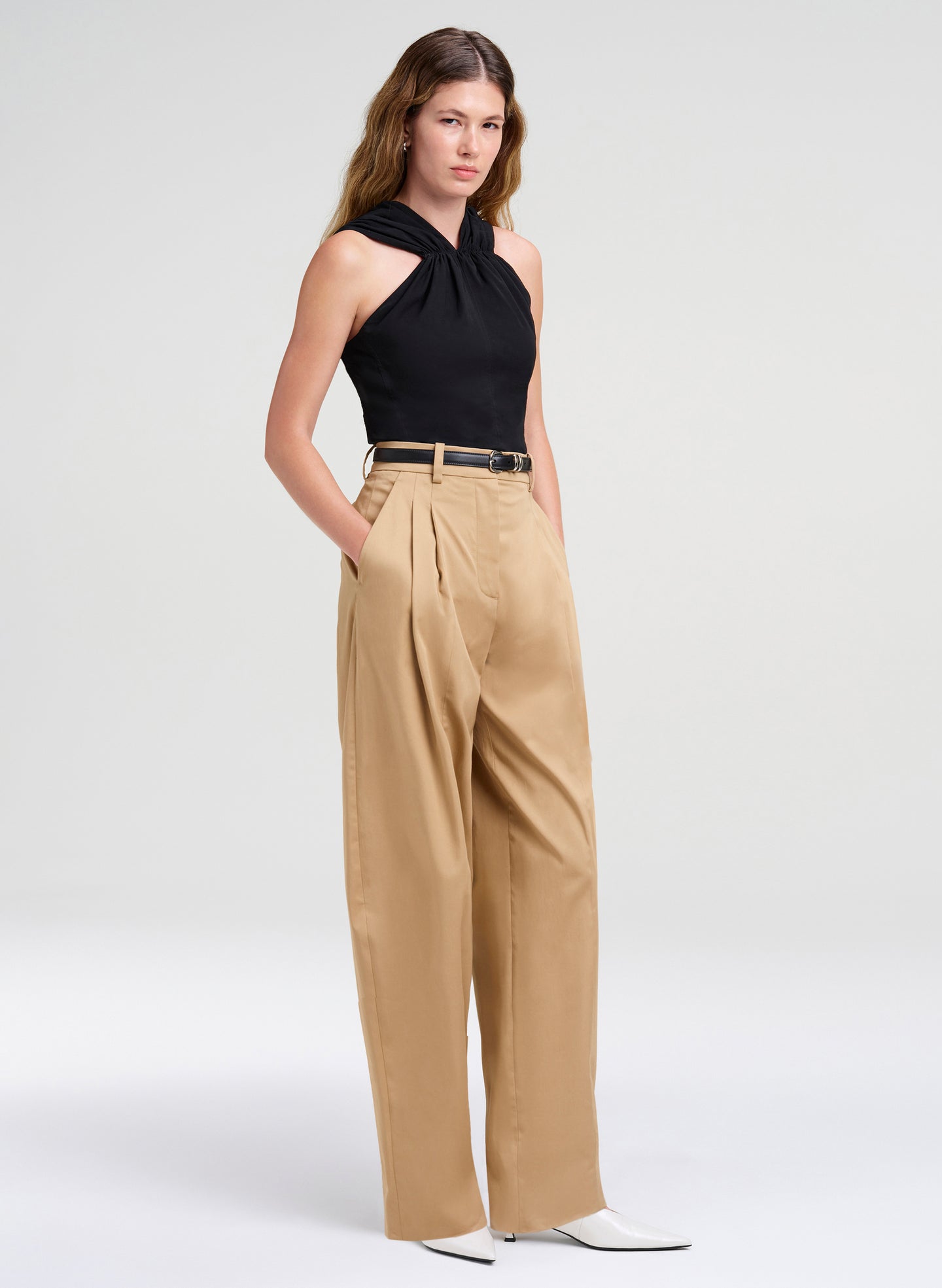 A model wearing a black halter top and high-waisted beige wide-leg trousers with a thin black belt, paired with white pointed-toe heels, against a light gray studio background.