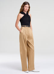 A model wearing a black halter top and high-waisted beige wide-leg trousers with a thin black belt, paired with white pointed-toe heels, against a light gray studio background.