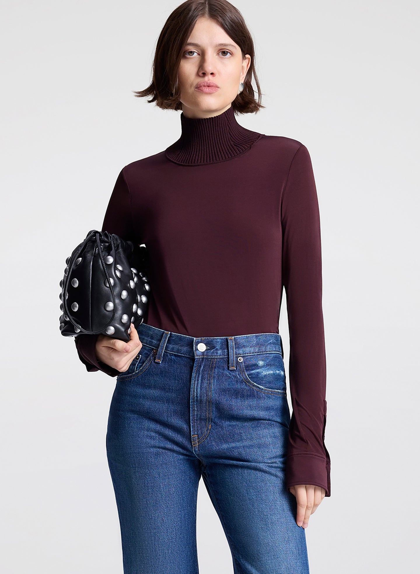 Woman wearing a dark purple turtleneck and blue jeans, holding a black studded handbag on a light gray background