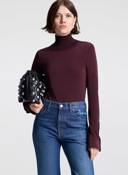 Woman wearing a dark purple turtleneck and blue jeans, holding a black studded handbag on a light gray background