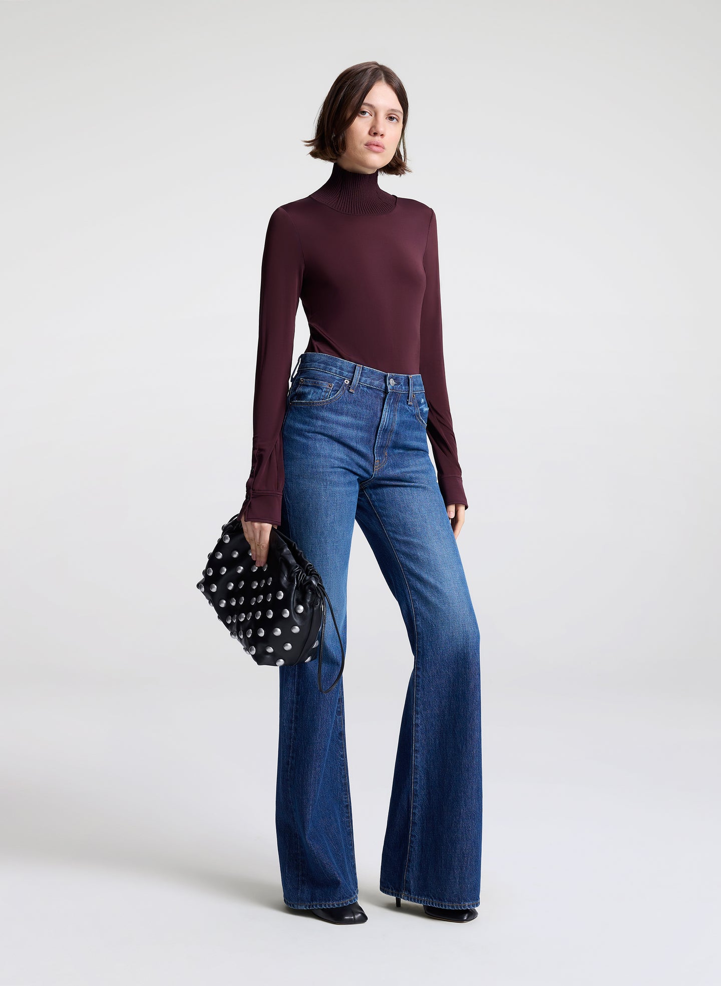 Woman wearing a dark purple turtleneck and blue jeans, holding a black clutch against a white background