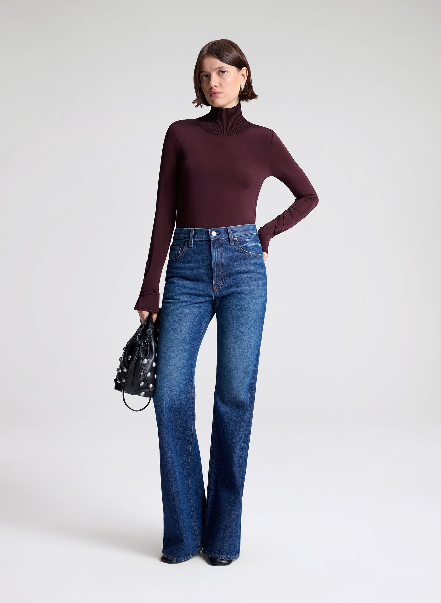 Woman wearing a dark turtleneck and blue jeans holding a black handbag on a white background