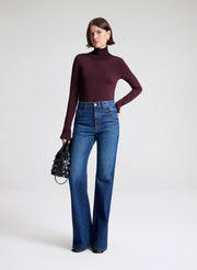 Woman wearing a dark turtleneck and blue jeans holding a black handbag on a white background
